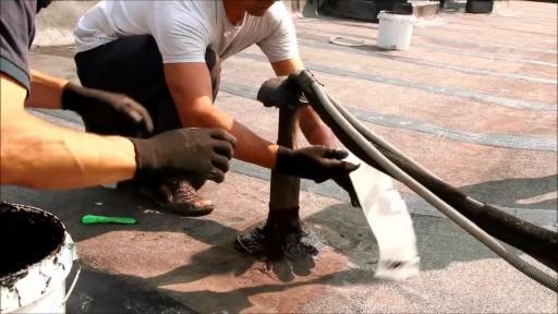 Dwóch mężczyzn w czarnych rękawicach uszczelnia dach płynną substancją z rury, obok stoi wiadro z czarną substancją i zielony nóż do cięcia papy.