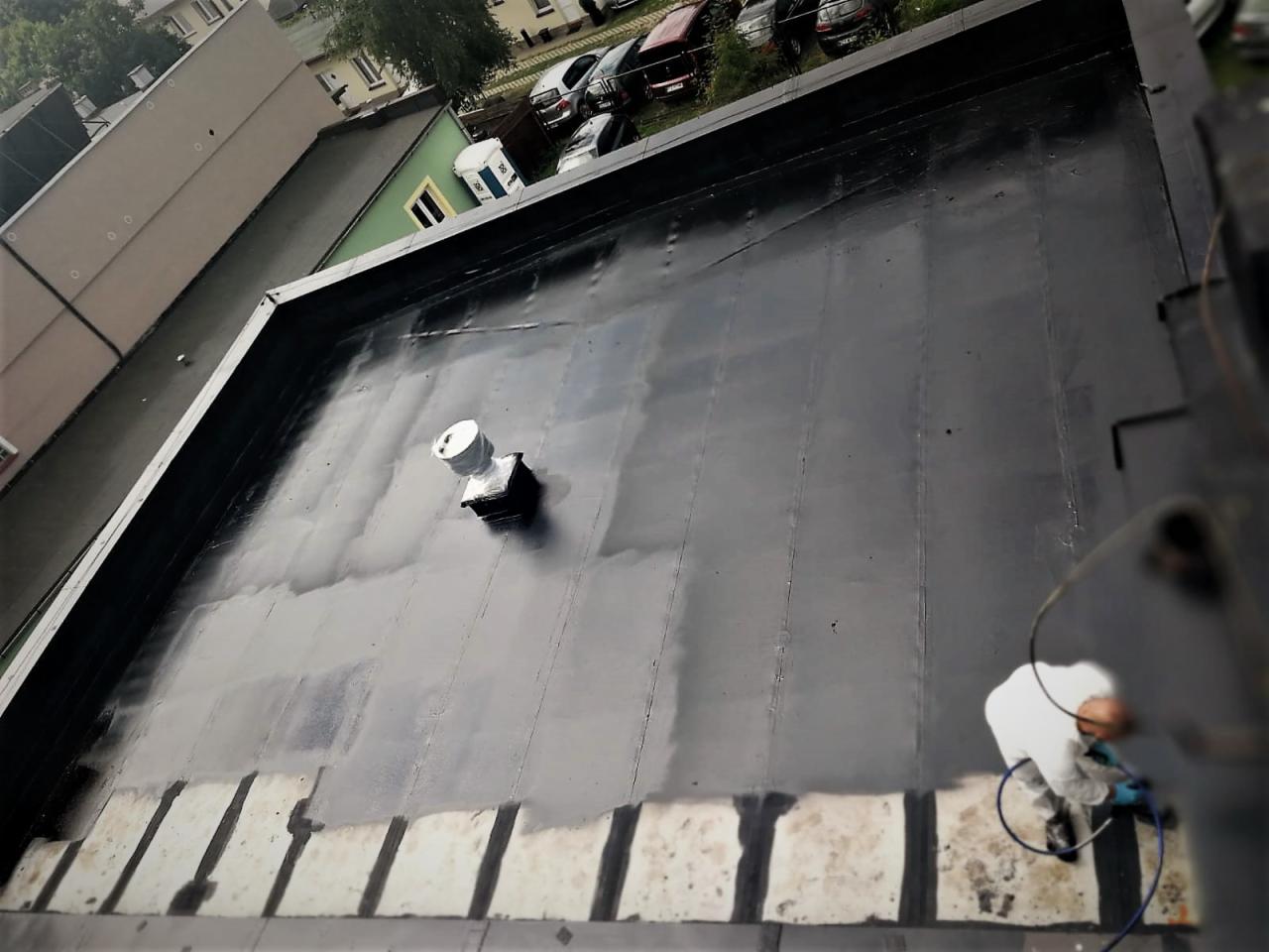 Wykończona izolacja płaskiego dachu budynku, widoczny pracownik w kombinezonie wykonujący ostatnie poprawki, elementy wentylacyjne oraz fragmenty niezabezpieczone izolacją.