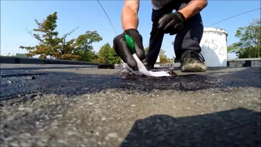 Pracownik w czarnych rękawicach aplikuje białą taśmę na czarną, bitumiczną powierzchnię dachu za pomocą zielonego narzędzia, obok stoi białe wiadro z napisem.