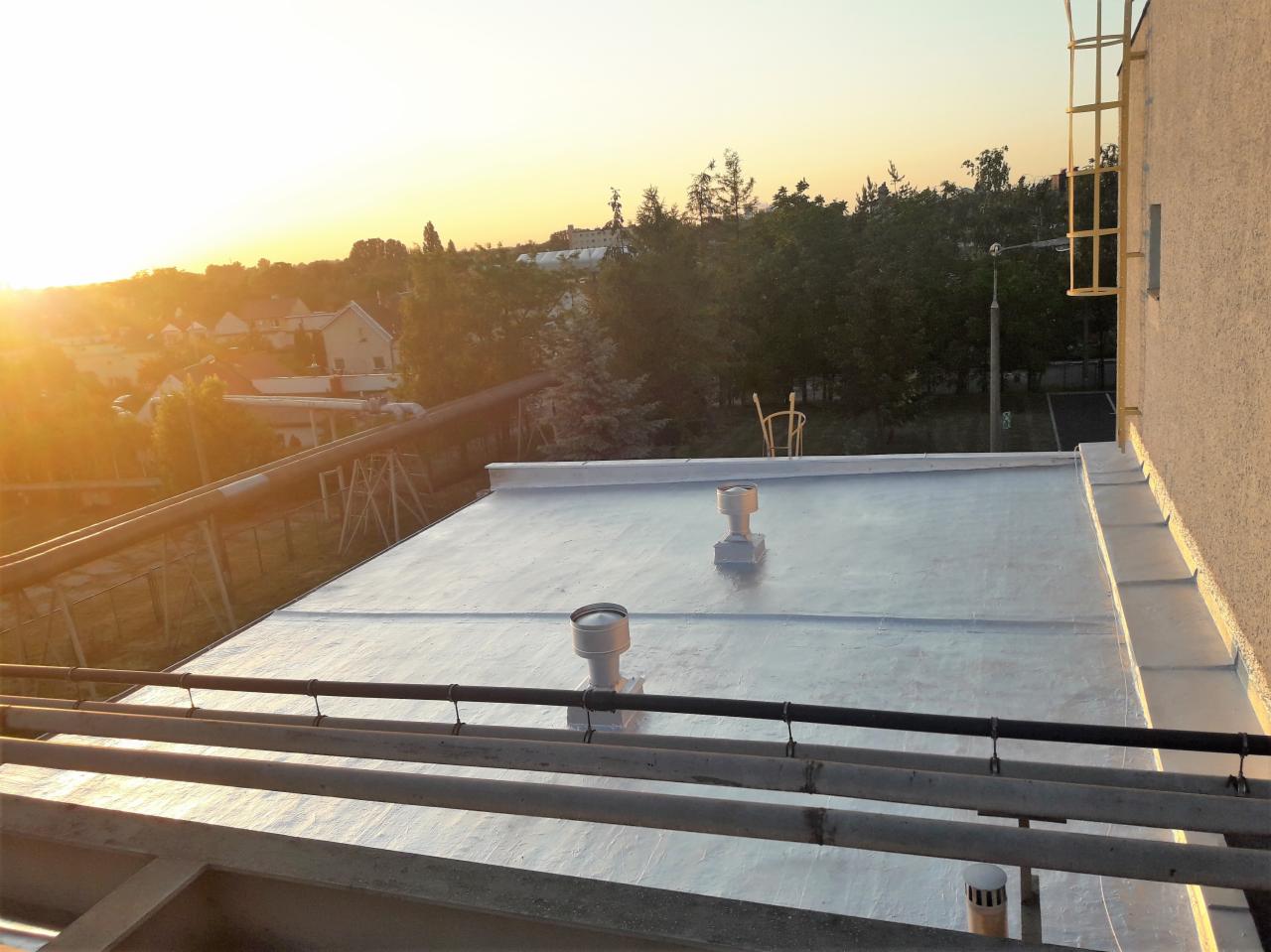 Jasnoszara, płaska powierzchnia dachu pokryta powłoką bitumiczną, widoczne dwa kominki wentylacyjne, rury i elementy konstrukcyjne dachu na tle zachodzącego słońca i zabudowań w oddali.