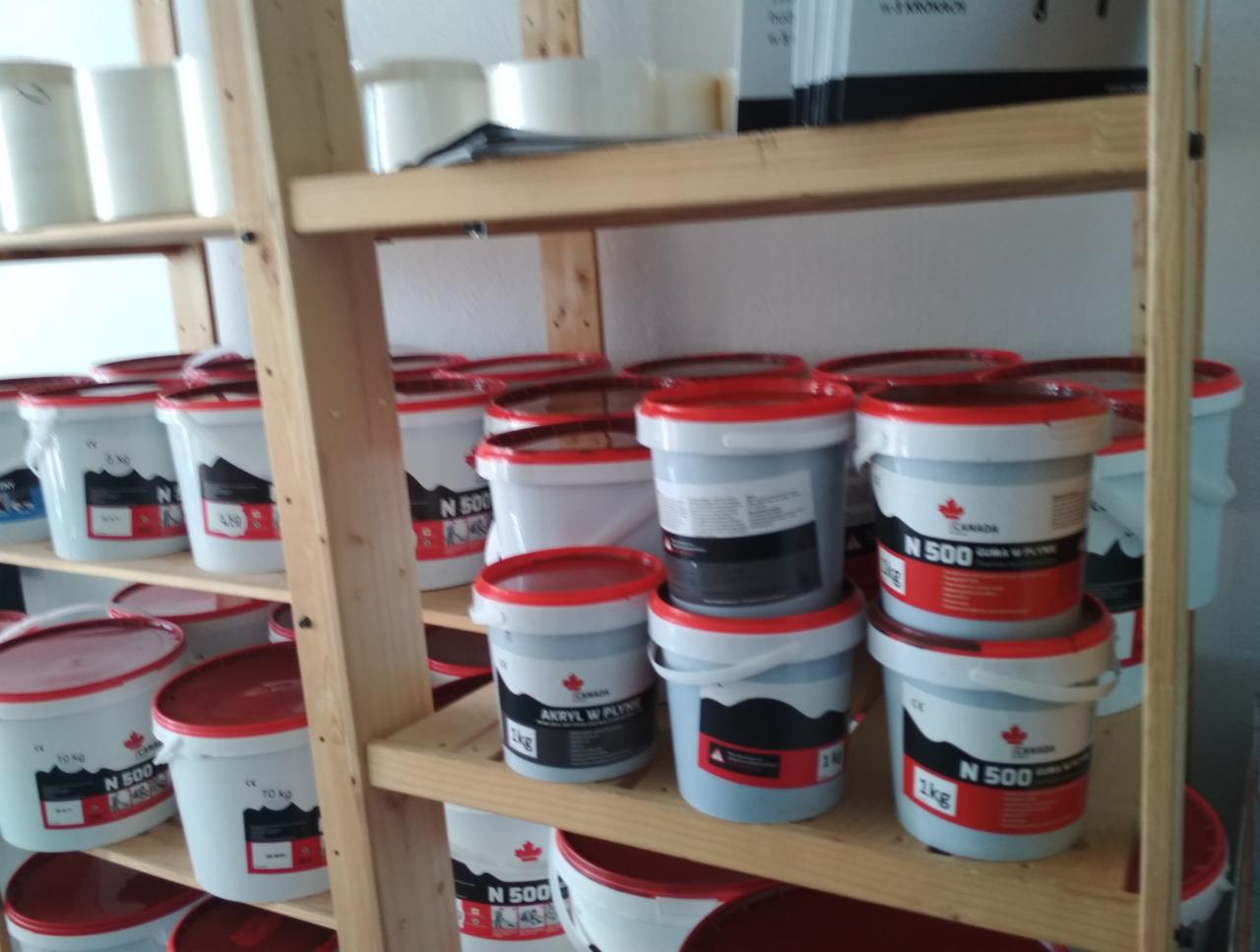 Półki z drewnianych desek wypełnione białymi wiaderkami z czerwonymi pokrywami, zawierającymi akryl w płynie i gumę w płynie, etykiety z logo Canada N500 widoczne na każdym wiaderku.