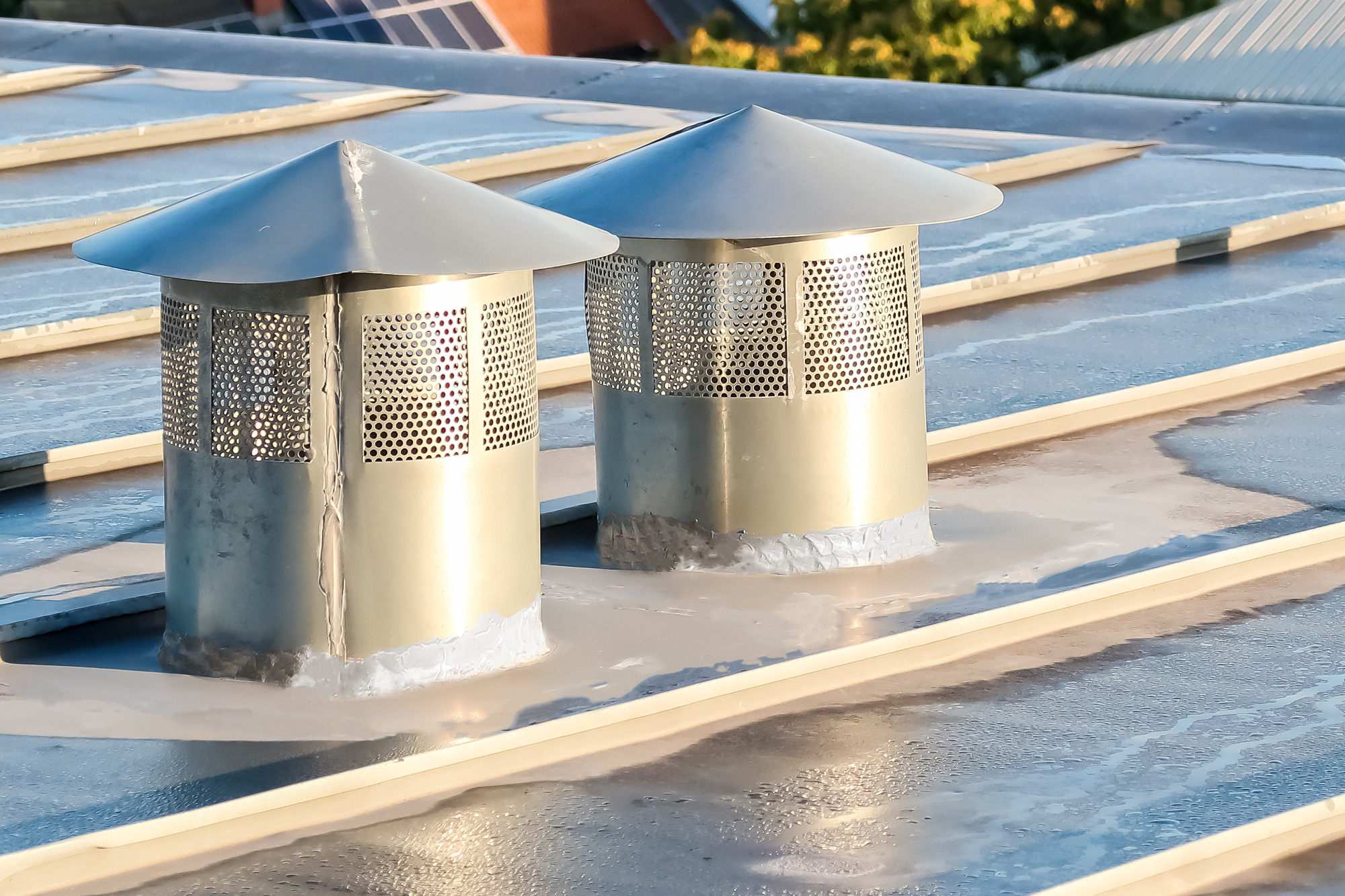 Dwa metalowe kominki wentylacyjne z perforowaną osłoną na blaszanym dachu z widocznymi panelami słonecznymi w tle.