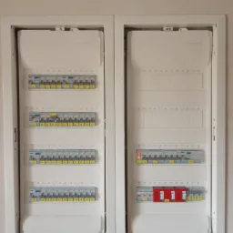 Dwa panele rozdzielcze w białej obudowie, z widocznymi bezpiecznikami i wyłącznikami, kilka z nich w pozycji wyłączonej (czerwone), zamontowane na jasnej ścianie.