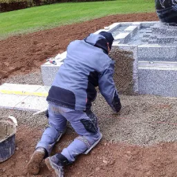 Pracownik w odzieży roboczej klęczy układając drobny żwir obok granitowej fontanny z motywem kuli ziemskiej, widoczny poziomica i wiadro z zaprawą.