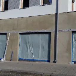 Elewacja budynku w trakcie prac: szpachlowana ściana z zabezpieczonymi folią i niebieską taśmą oknami, drabina aluminiowa i poziomica oparta o ścianę.