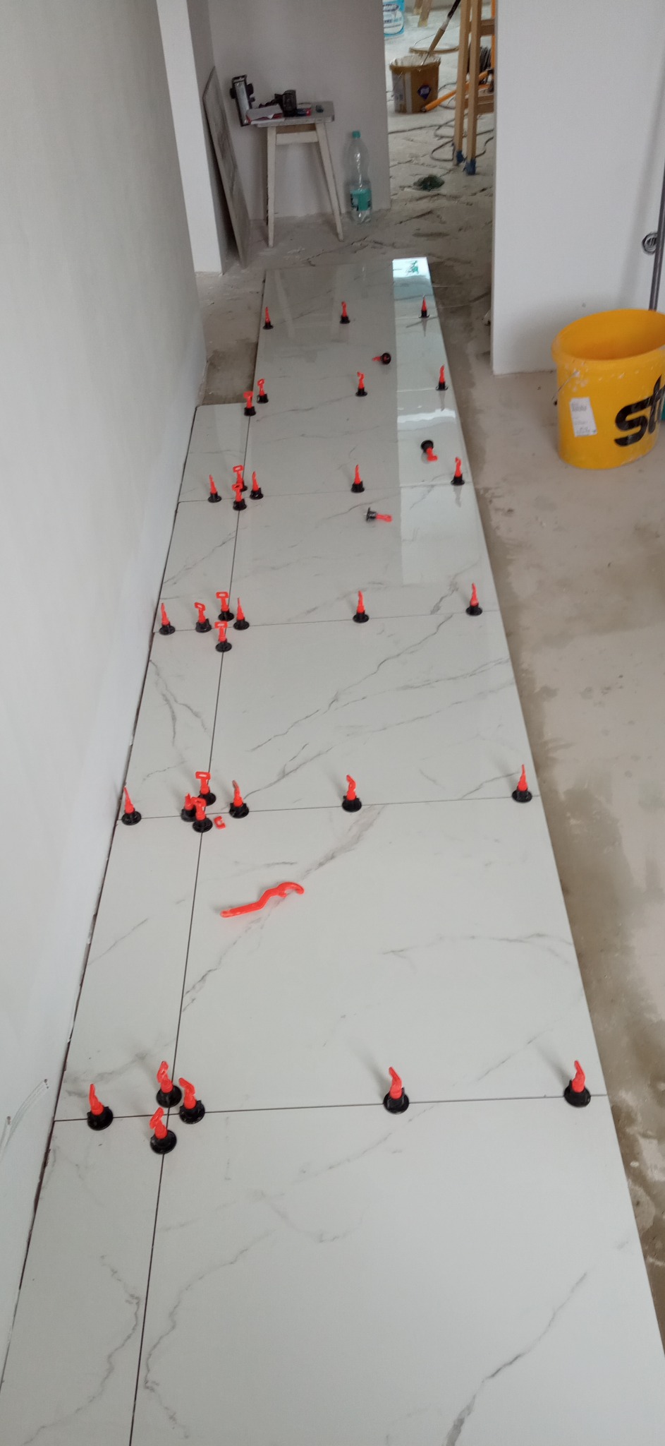 Układanie białych płytek podłogowych z marmurowym wzorem, z użyciem czerwonych klinów poziomujących i krzyżyków dystansowych; widoczny fragment żółtego wiadra z logo producenta.