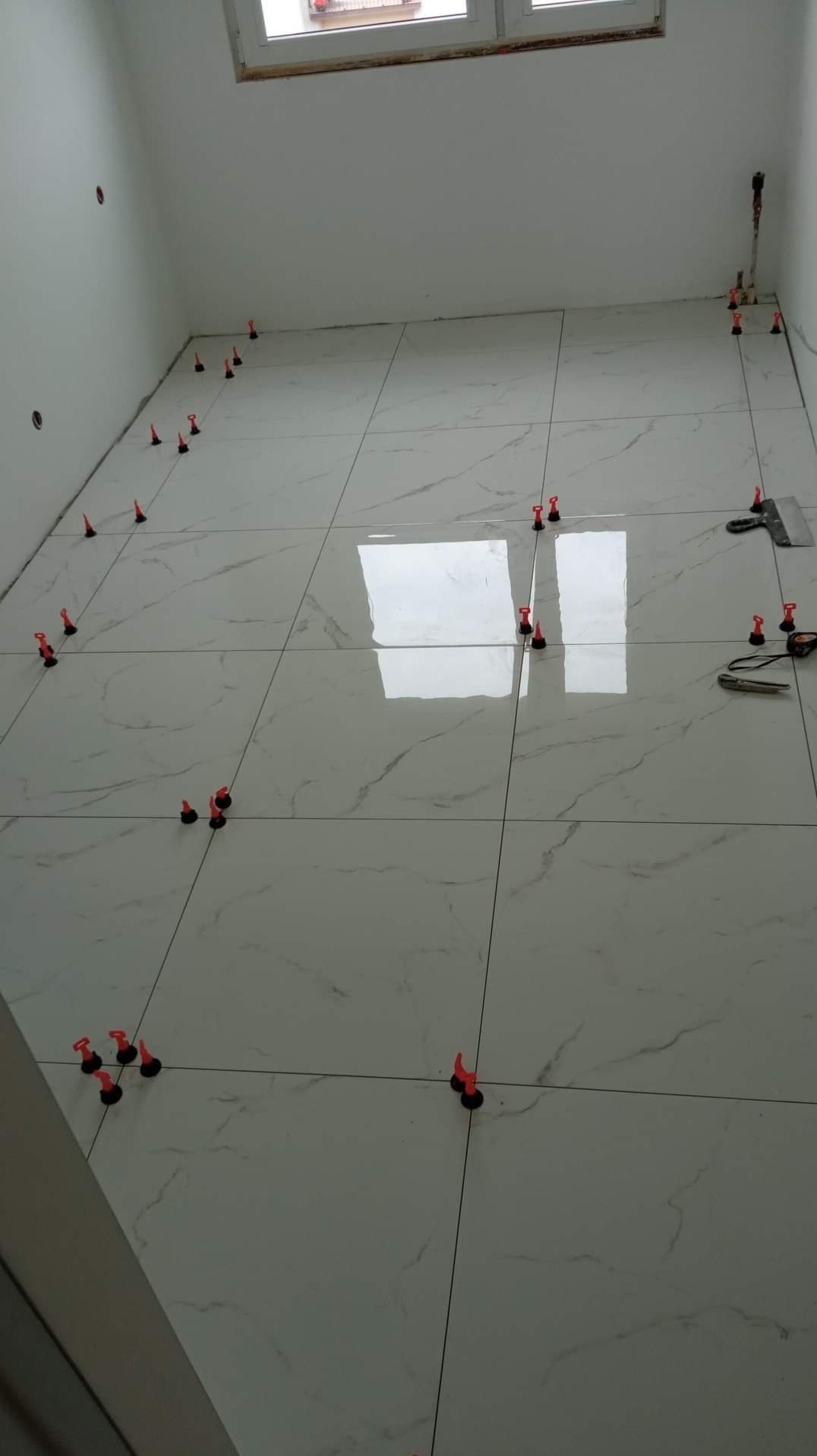 Układanie białych płytek podłogowych z marmurowym wzorem, widoczne czerwone kliny poziomujące, szpachelka i miarka leżące na podłodze, jasne wnętrze łazienki w trakcie remontu.