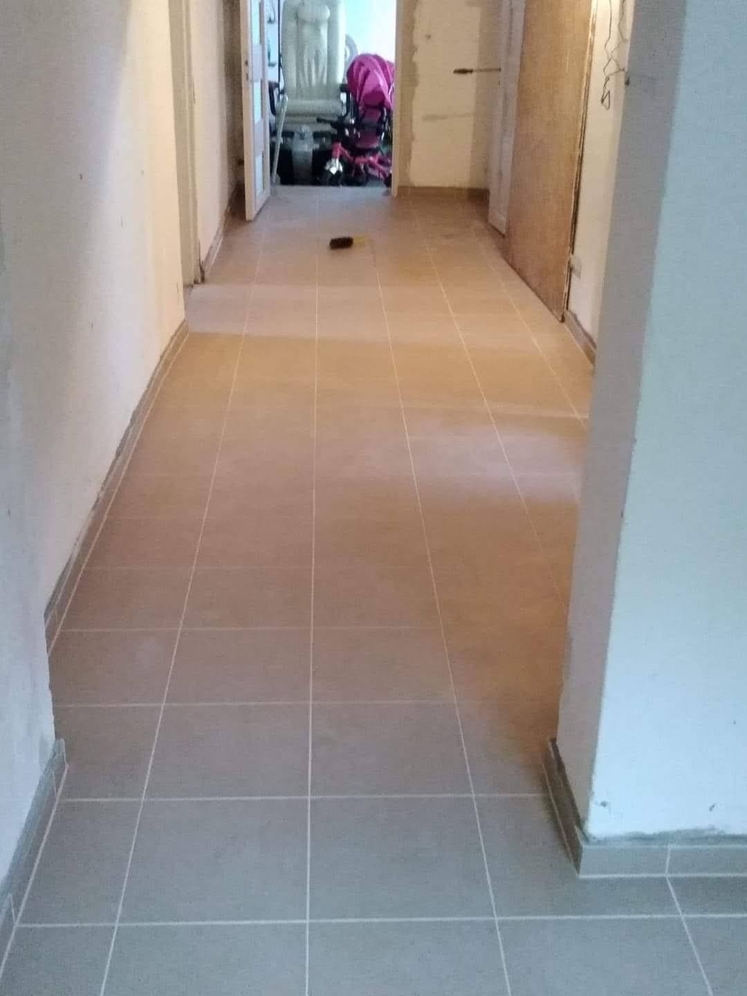 Wyłożona jasnymi, kwadratowymi płytkami podłoga korytarza w trakcie remontu, widoczne niedokończone ściany i otwarte drzwi do innych pomieszczeń.