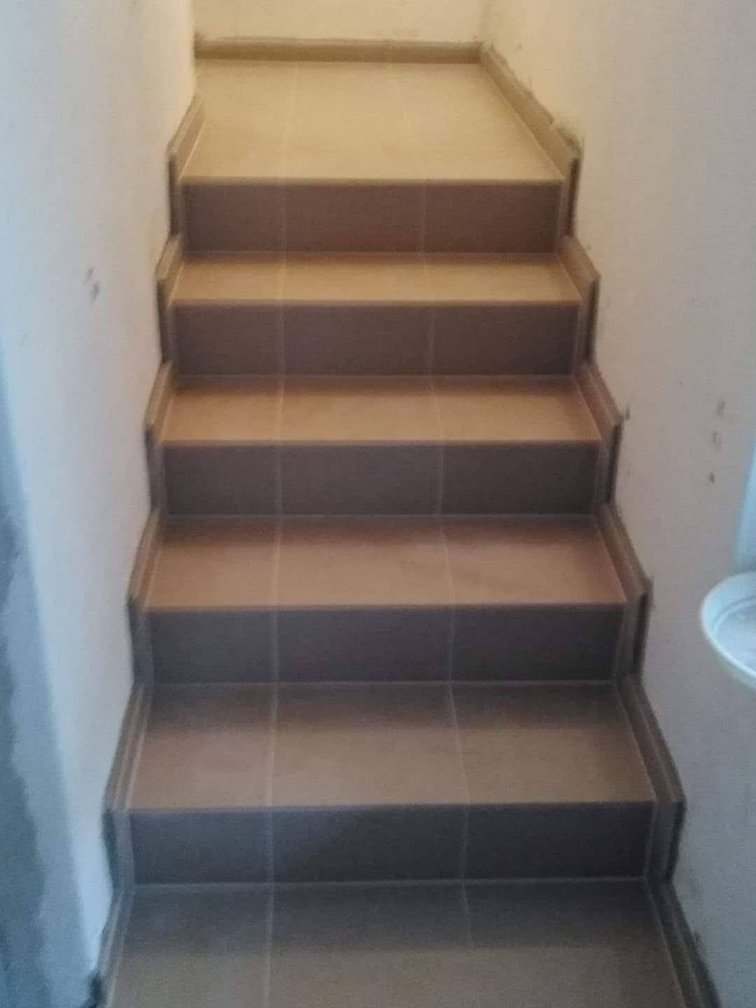 Schody wyłożone brązowymi płytkami ceramicznymi, widok z góry, fuga w kolorze płytek, ściany pomalowane na biało, widoczne niedoskonałości na ścianie.