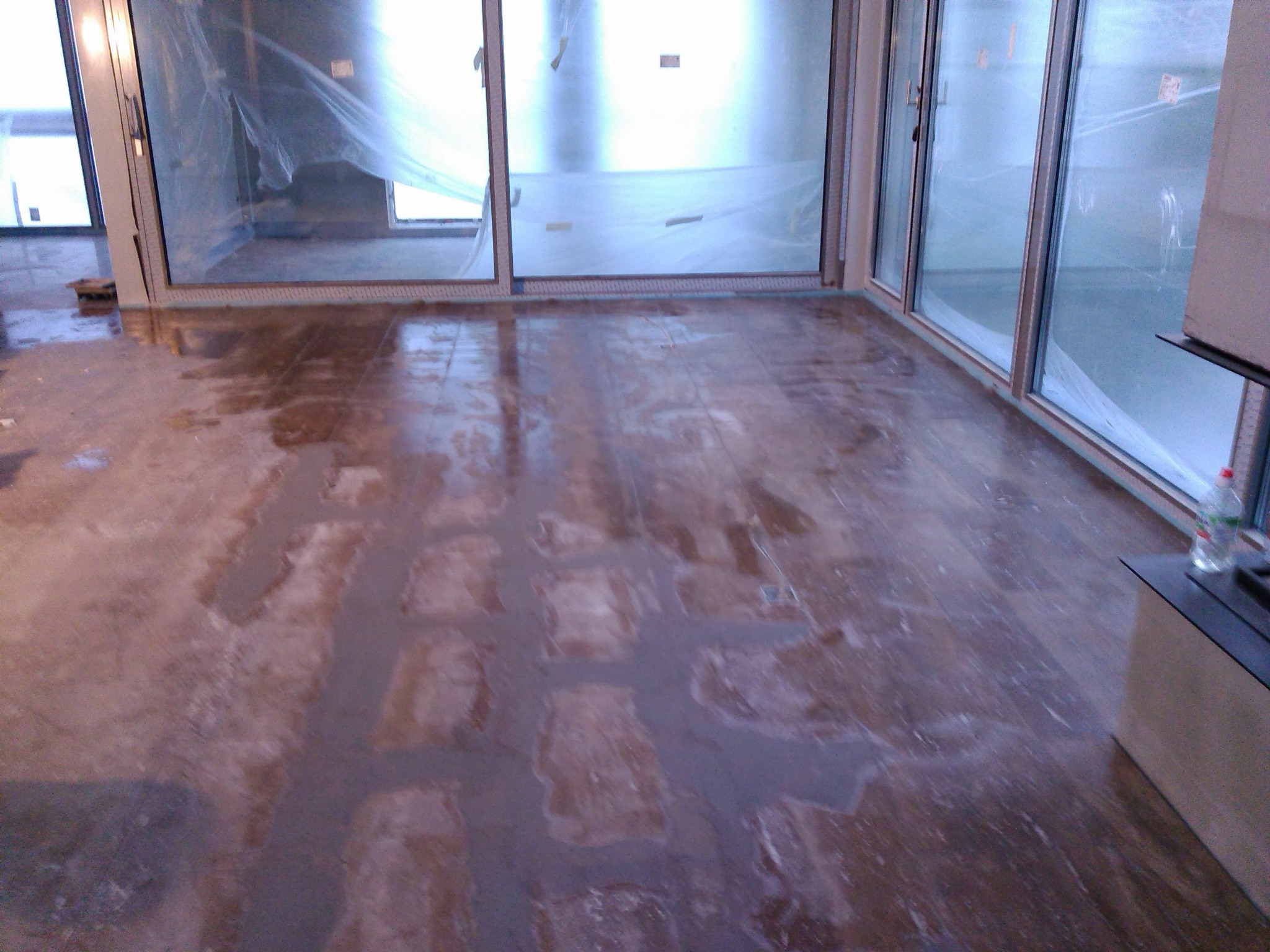 Wnętrze w trakcie prac przygotowawczych do wykończenia podłogi, widoczne duże okna zabezpieczone folią malarską i częściowo nałożona wylewka wyrównująca na drewnianej podłodze.