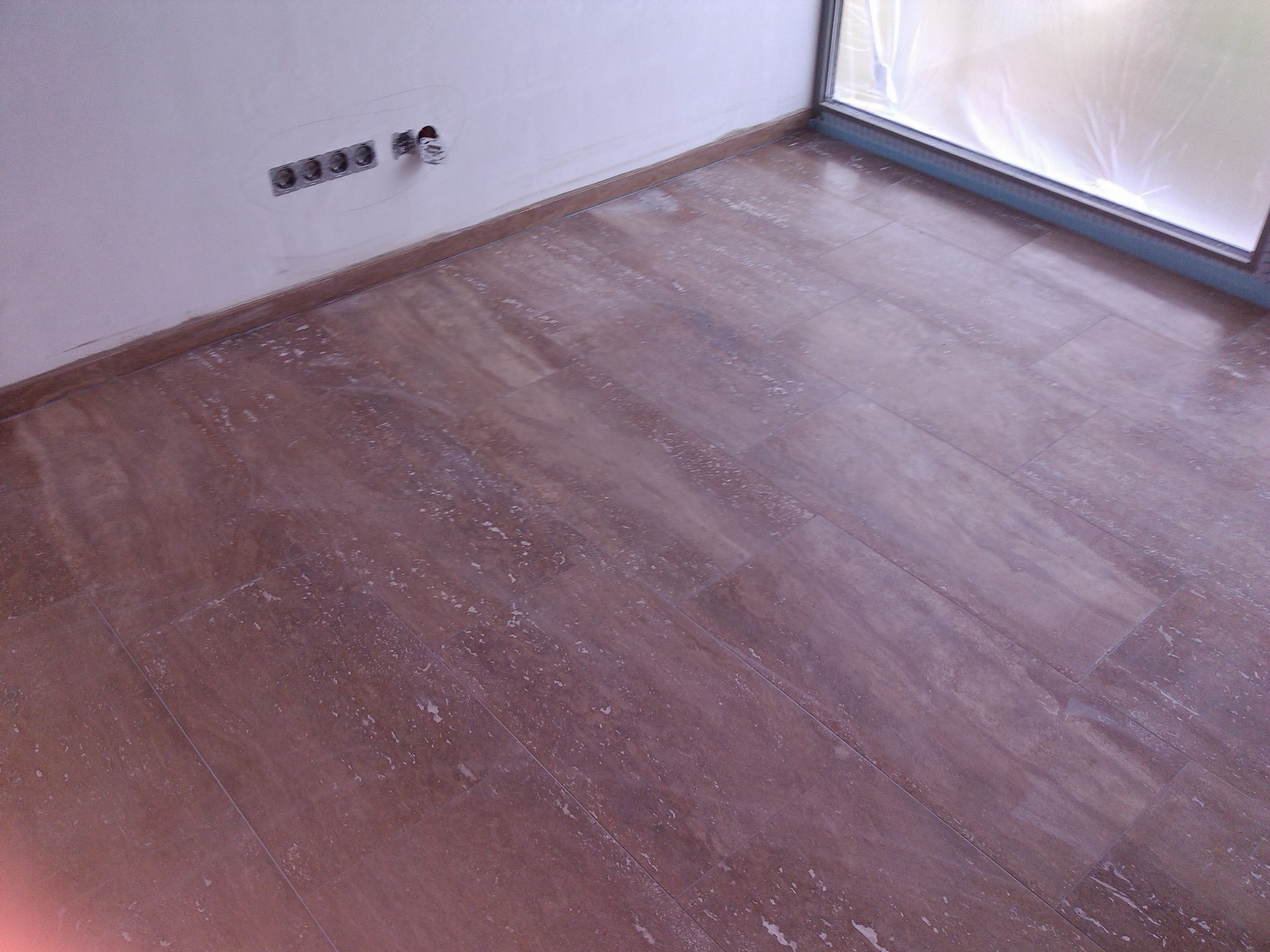 Podłoga wyłożona brązowymi płytkami imitującymi drewno, ściana z gniazdkami elektrycznymi przed malowaniem, okno zabezpieczone folią.