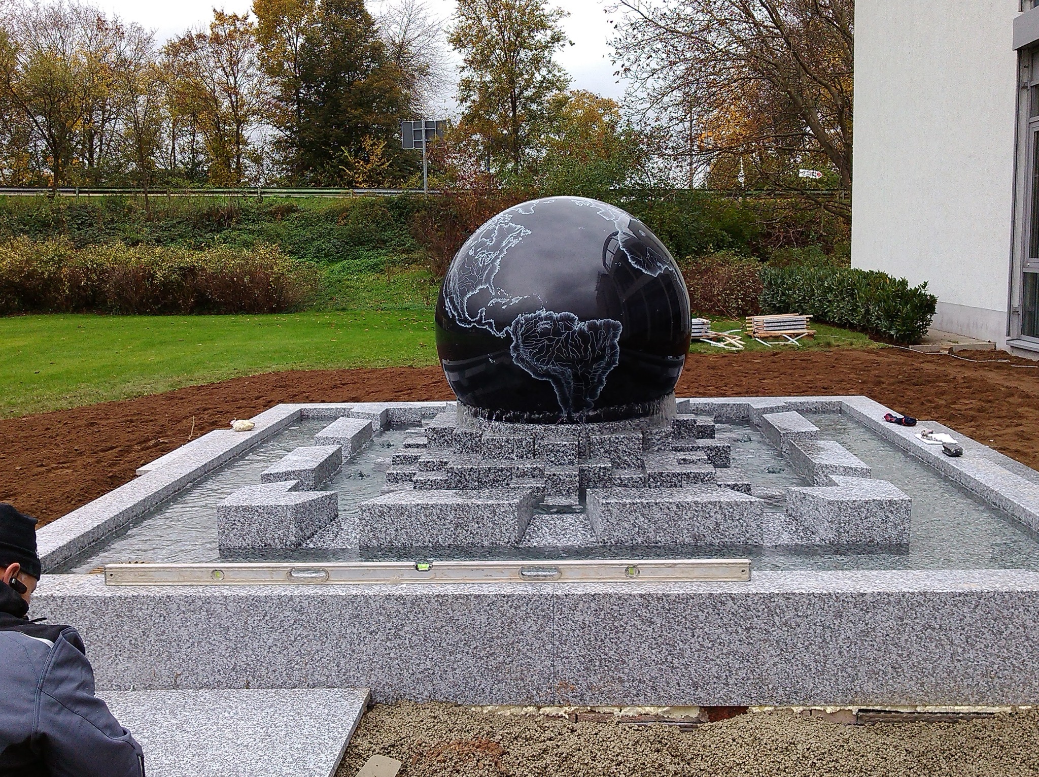 Czarna kula z wygrawerowaną mapą świata, umieszczona w nowoczesnej, granitowej fontannie z kaskadami wodnymi, otoczona trawnikiem i budynkiem w tle.