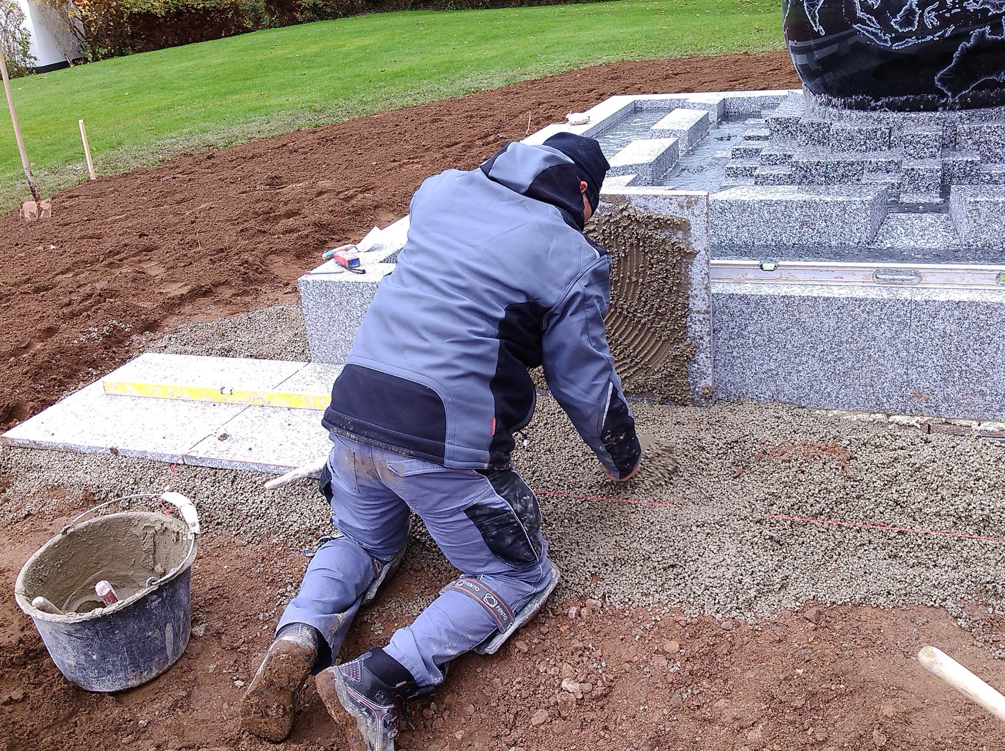Pracownik w odzieży roboczej klęczy układając drobny żwir obok granitowej fontanny z motywem kuli ziemskiej, widoczny poziomica i wiadro z zaprawą.