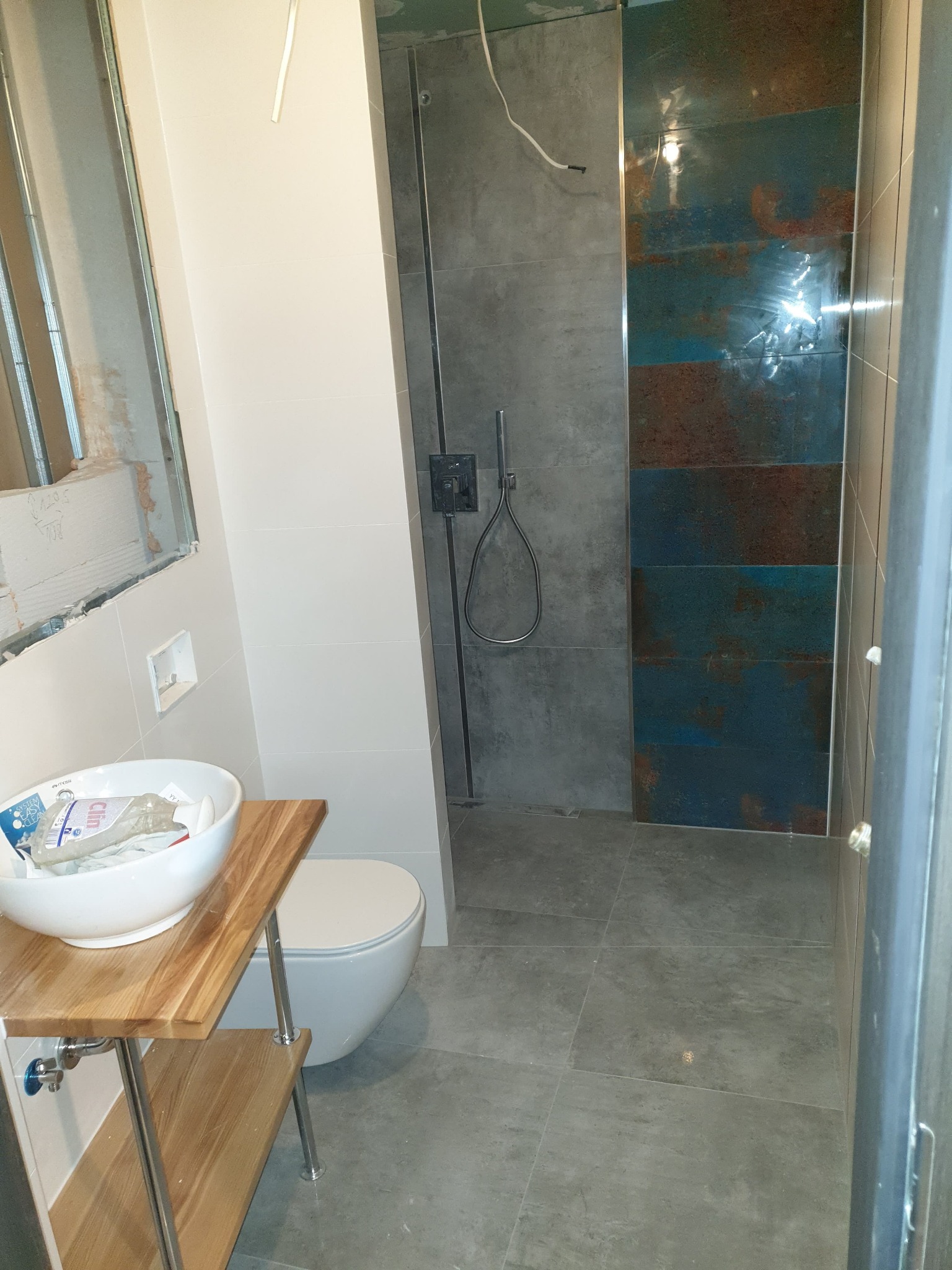 Łazienka w trakcie remontu: widoczne fragmenty ściany z odsłoniętą konstrukcją, umywalka na drewnianym stojaku, toaleta oraz kabina prysznicowa z płytkami imitującymi rdzę.