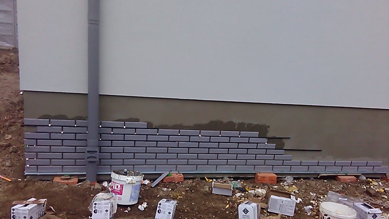 Montaż elewacji z szarej cegły klinkierowej na fundamencie domu, widoczne puszki i narzędzia budowlane na ziemi.