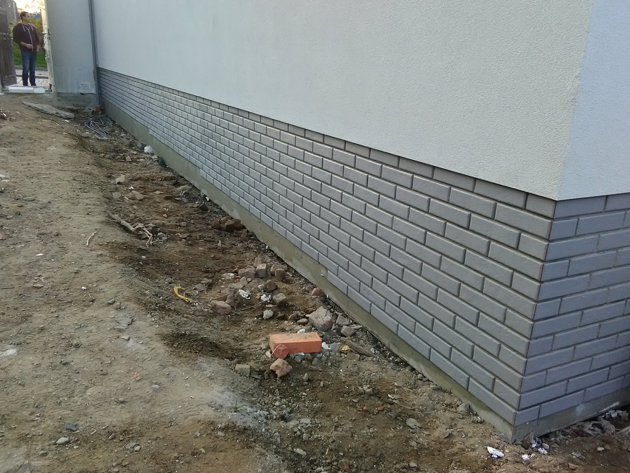 Wykończenie elewacji budynku płytkami imitującymi cegłę w szarym odcieniu, widoczny fragment ściany i narożnik, teren wokół w trakcie prac ziemnych.
