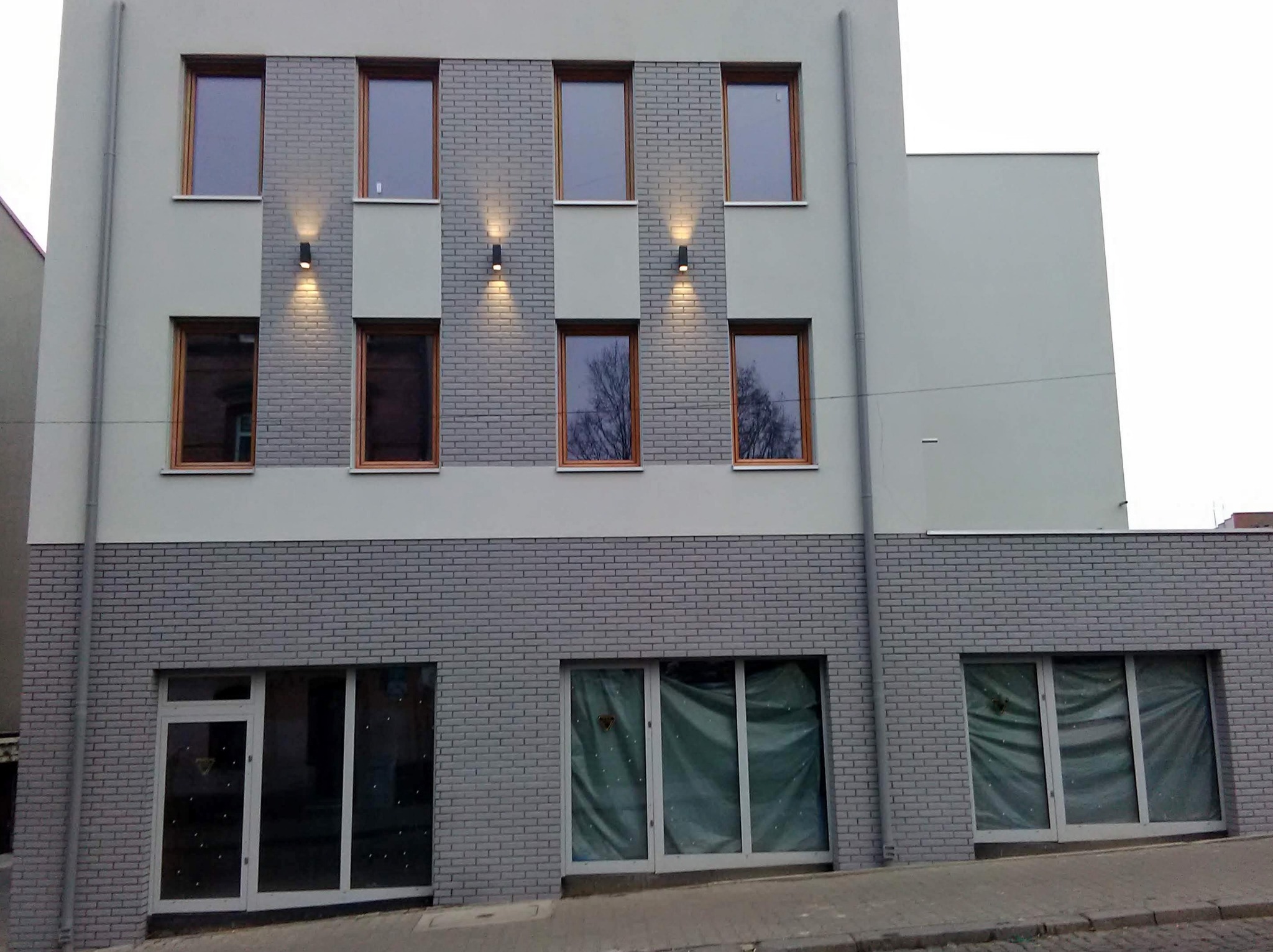 Fasada budynku po remoncie z szarą cegłą klinkierową na dole, drewnianymi oknami i nowoczesnym oświetleniem zewnętrznym.