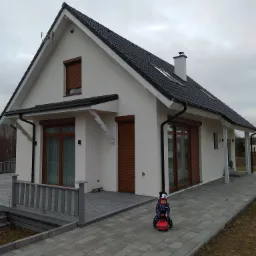 Nowoczesny dom jednorodzinny z białymi ścianami, brązowymi roletami i czarnym dachem, z dzieckiem na czerwonym jeździku na kostce brukowej.