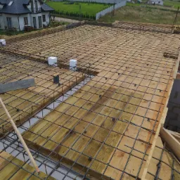 Przygotowanie szalunku i zbrojenia pod wylewkę fundamentu na płycie, widoczne drewniane deski, stalowe pręty zbrojeniowe tworzące siatkę, białe kostki dystansowe, w tle zabudowa jednorodzinna.