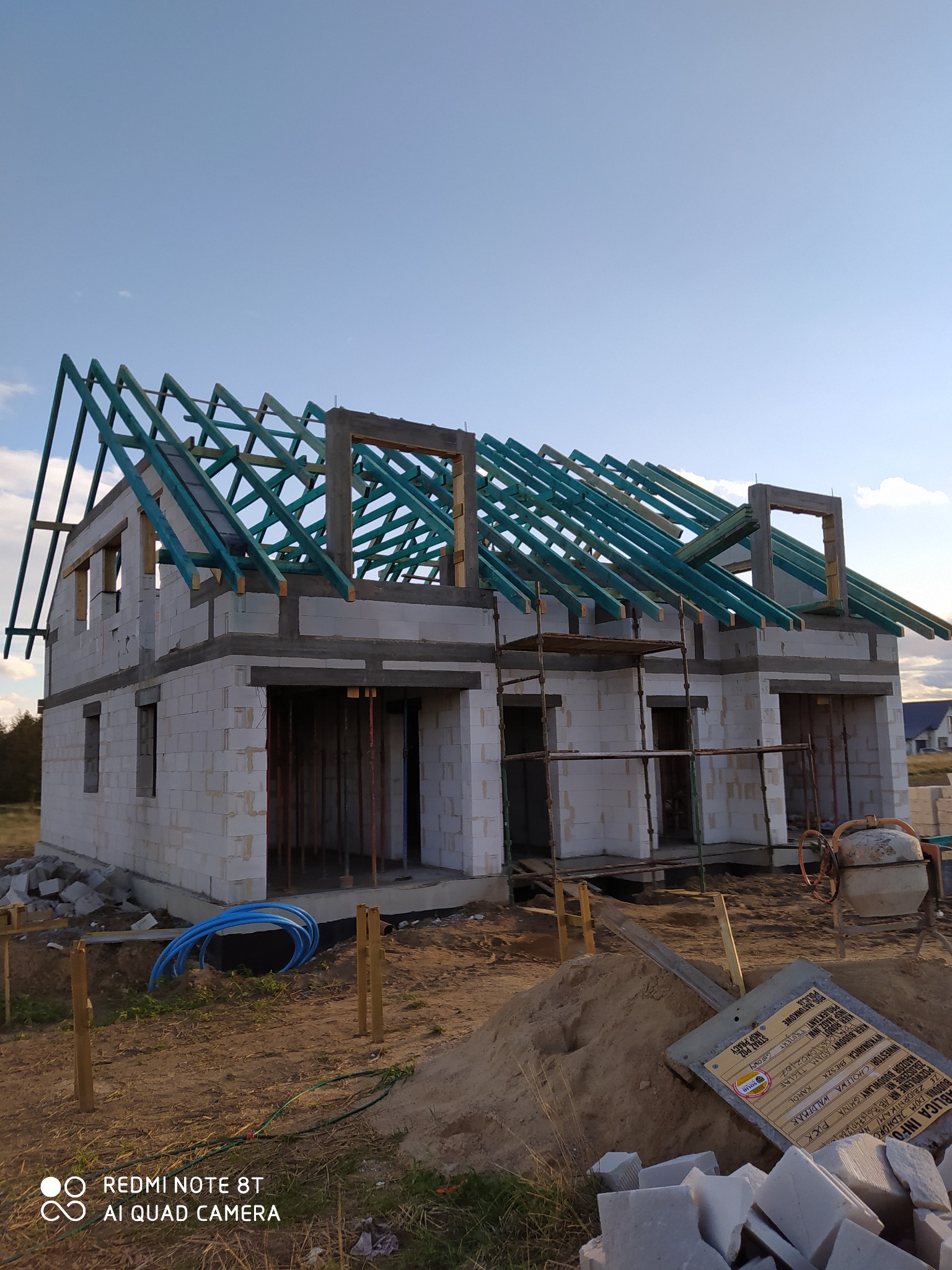 Budowa domu jednorodzinnego: wzniesione ściany z bloczków i konstrukcja dachu z impregnowanego drewna na tle błękitnego nieba, widoczne rusztowania i materiały budowlane.