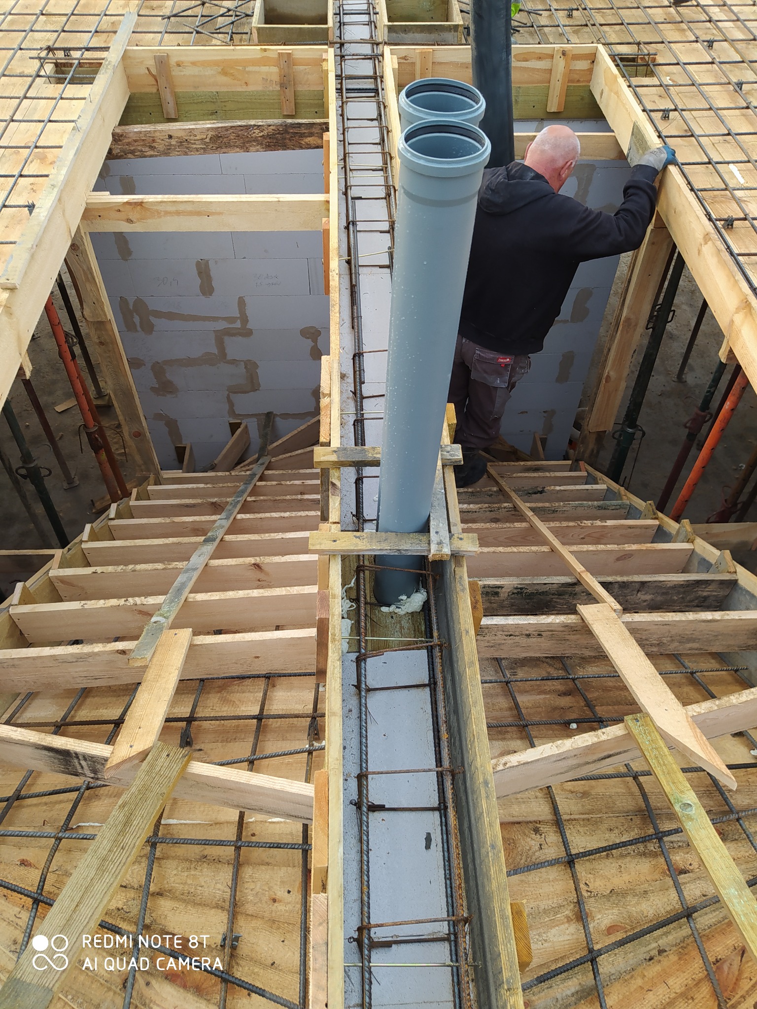 Konstrukcja szalunku zbrojonego na wylewkę fundamentu, widoczny pracownik w rękawicach montujący rury kanalizacyjne, ściany z bloczków betonowych w tle.