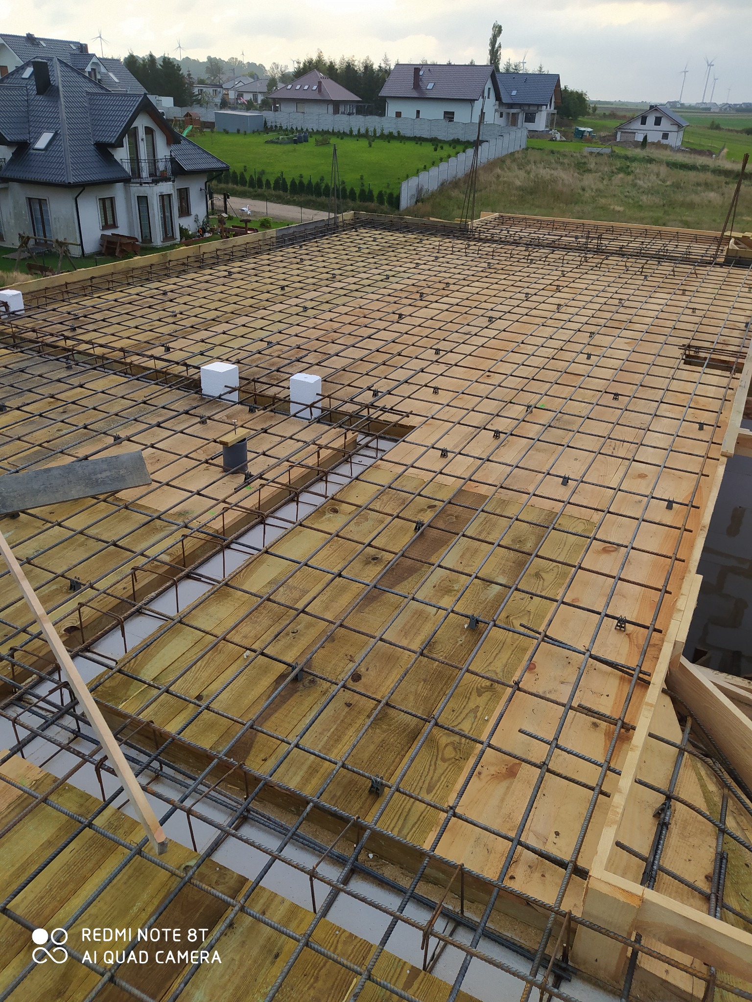 Przygotowanie szalunku i zbrojenia pod wylewkę fundamentu na płycie, widoczne drewniane deski, stalowe pręty zbrojeniowe tworzące siatkę, białe kostki dystansowe, w tle zabudowa jednorodzinna.