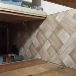 Ściana wykończona panelami ściennymi imitującymi drewno ułożonymi w jodełkę, widok z dołu, fragment szafki i akcesoria budowlane w tle.