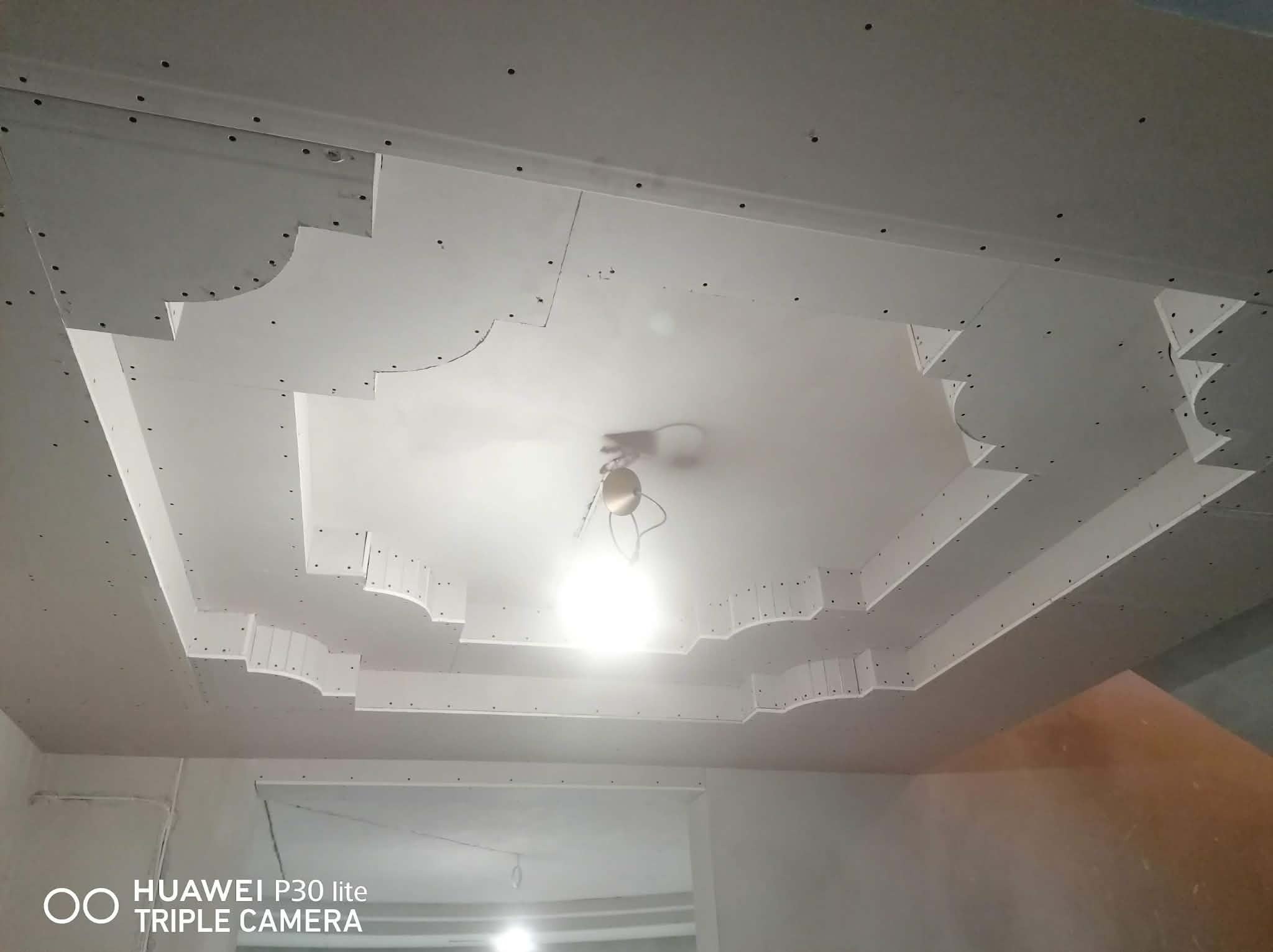 Dekoracyjny sufit z płyt kartonowo-gipsowych z oświetleniem centralnym, widoczne ślady po wkrętach, surowy stan wykończenia.