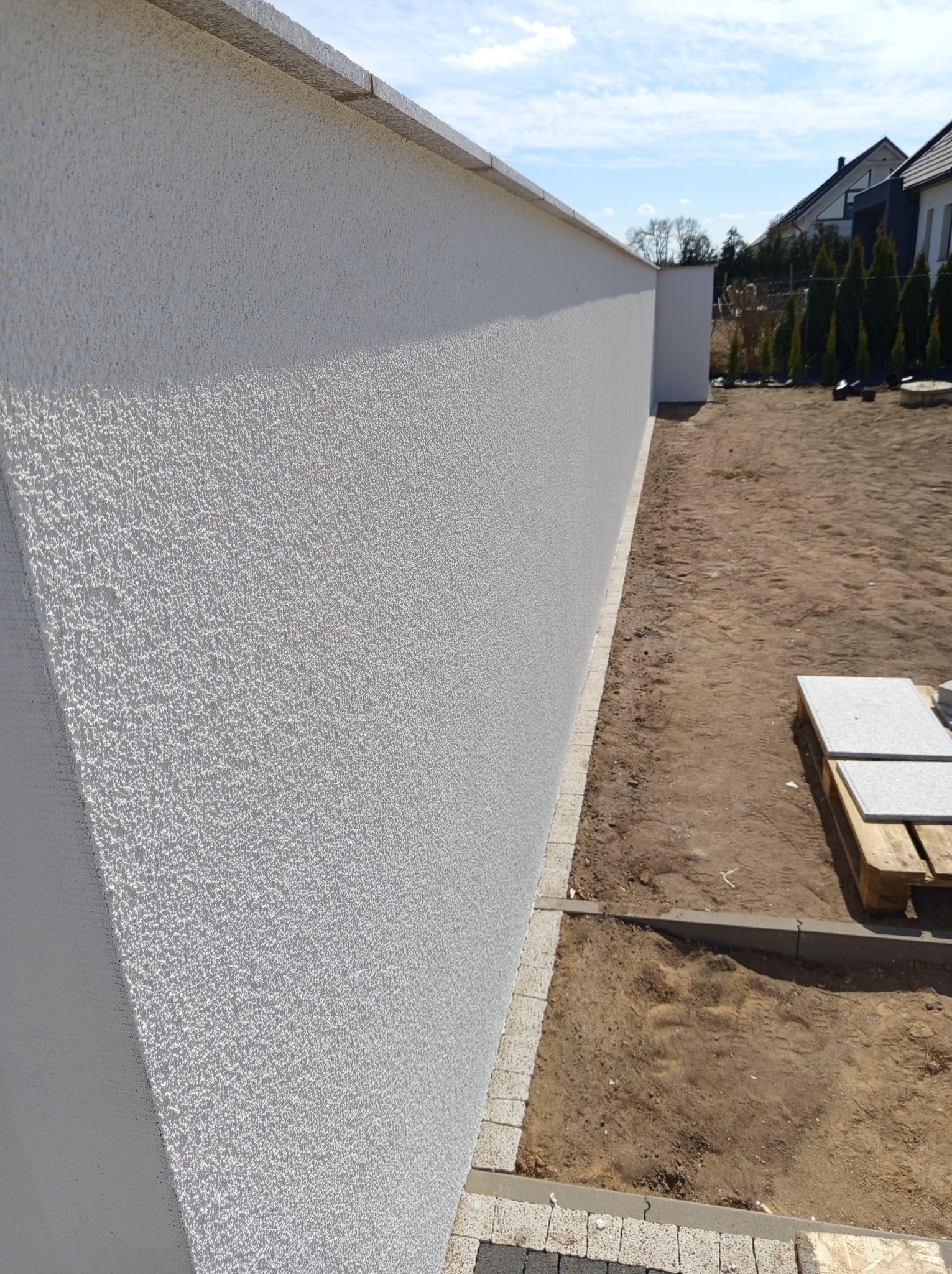 Biały, teksturowany mur ogrodzeniowy z betonową czapą, widziany z perspektywy bocznej. W tle domy i drzewa. Na ziemi paleta z płytami.