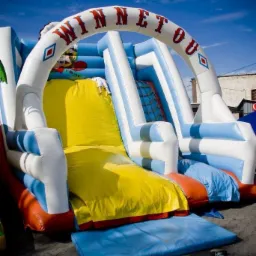 Niski kąt ujęcia na dmuchaną zjeżdżalnię w kolorach niebieskim, białym, żółtym i pomarańczowym z napisem 'WINNETOU' na górze, na tle błękitnego nieba.
