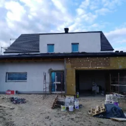 Dom jednorodzinny w trakcie ocieplania, widoczne rusztowania, styropian, puszki z farbą i paczki wełny mineralnej Rockwool, okna zabezpieczone folią, częściowo otynkowana ściana.