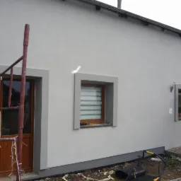 Świeżo otynkowana elewacja domu jednorodzinnego w odcieniach szarości, z widocznymi ramami okiennymi i drzwiami wejściowymi, fragment rusztowania po lewej stronie oraz kosiarka do trawy w dolnej...