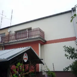 Dwukondygnacyjny budynek po renowacji elewacji, z białymi ścianami, brązowym pasem i balkonem z brązową balustradą, widoczne anteny na dachu i zadaszenie z kwiatami w ogrodzie.