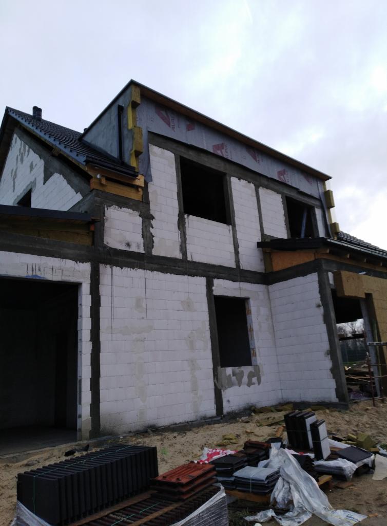 Dom w trakcie ocieplania elewacji, widoczne białe bloczki, częściowo ocieplone ściany, przygotowane dachówki na ziemi.