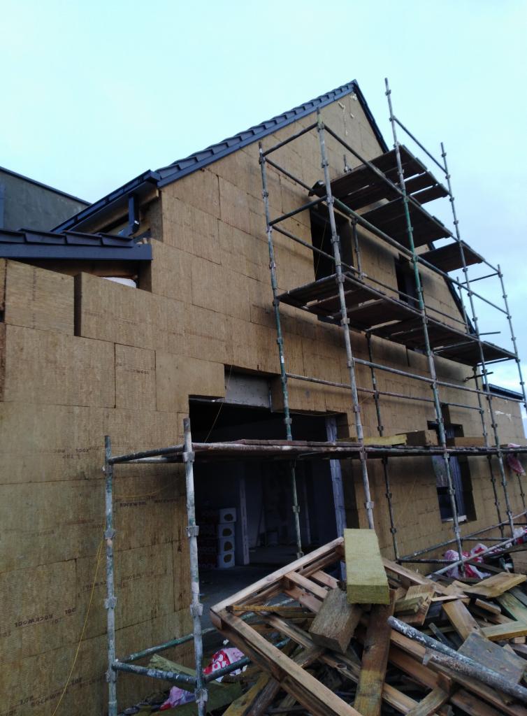 Dom w trakcie ocieplania, widoczne rusztowania i materiał izolacyjny na ścianach zewnętrznych, stos drewnianych odpadów na pierwszym planie.