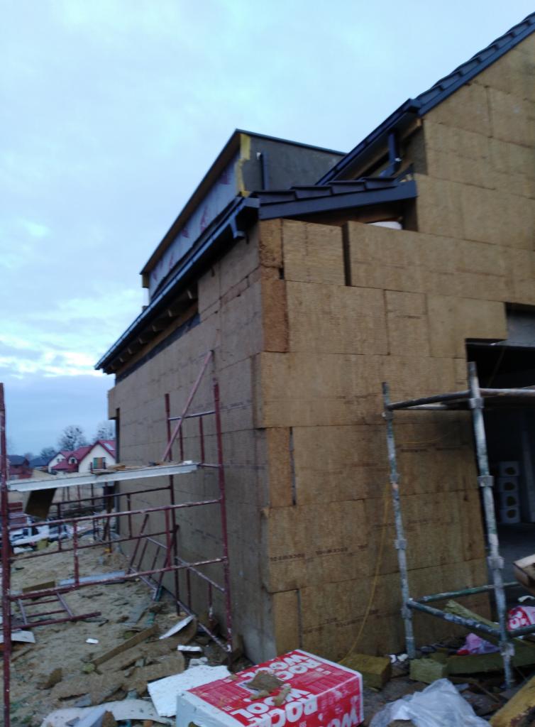 Budynek w trakcie ocieplania, widoczne płyty izolacyjne na elewacji, rusztowania i materiały budowlane na ziemi, pochmurne niebo w tle.