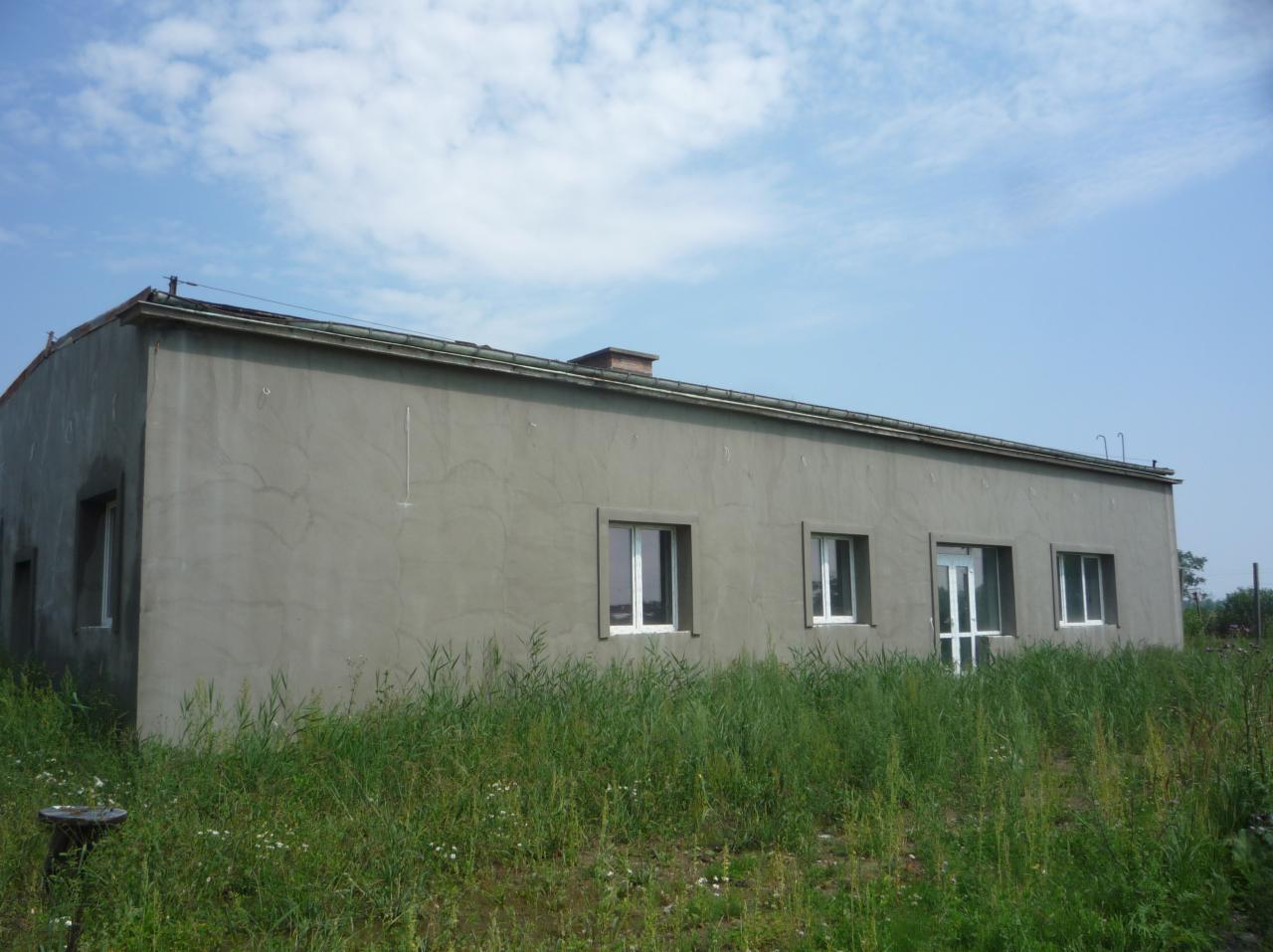 Surowa elewacja budynku jednokondygnacyjnego z widoczną fakturą, otoczona wysoką trawą i chwastami, z fragmentem nieba w tle.