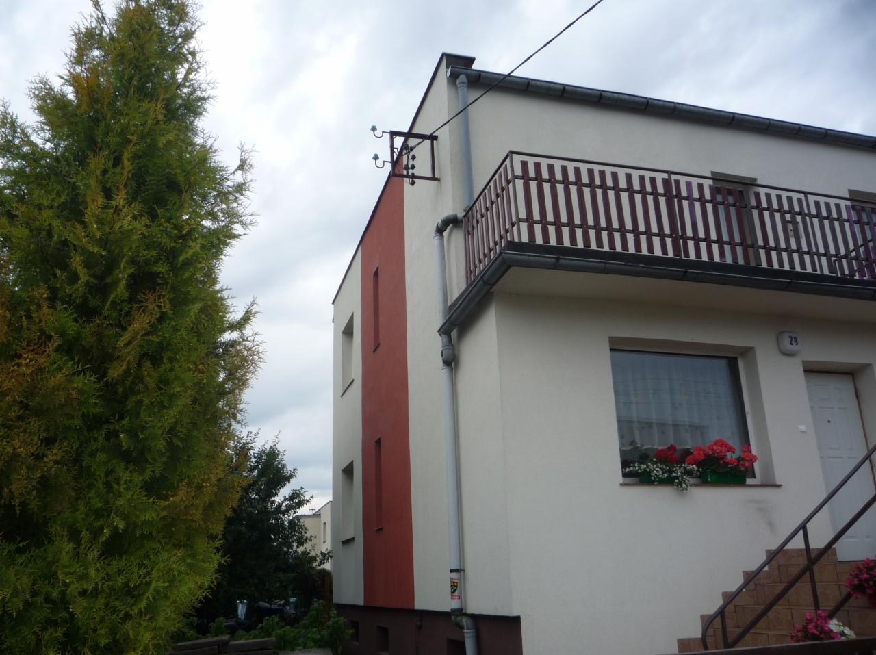 Dwukondygnacyjny dom z balkonem i kwiatami w oknie, z fragmentem elewacji w kolorze czerwonym, widok z poziomu gruntu, obok wysoki tuja.