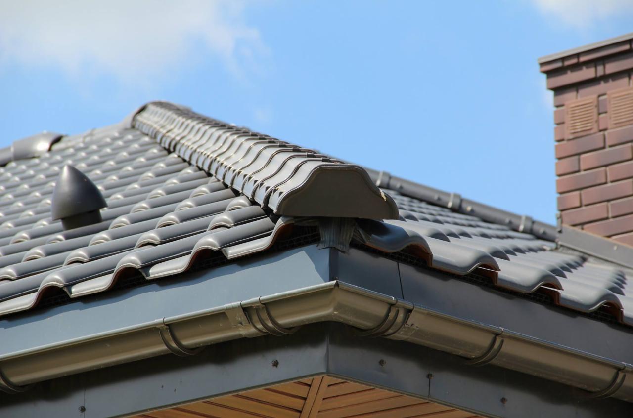 Szczegółowe ujęcie dachu z ciemnoszarej dachówki ceramicznej Titania, z widocznymi elementami wentylacji i systemem rynnowym, na tle błękitnego nieba i komina z cegły.