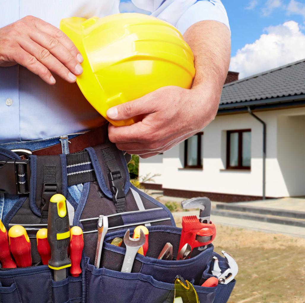 Budowlaniec trzyma żółty kask, w pasie narzędziowym widoczne różne narzędzia, w tle biały dom jednorodzinny z ciemną dachówką.