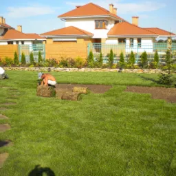 Dwaj mężczyźni rozkładają rolki trawy na przygotowanym gruncie w ogrodzie przed domem z czerwonym dachem; widoczna ścieżka z kamieni prowadząca przez trawnik i ozdobne tuje wzdłuż płotu.