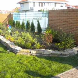 Kaskadowa rabata kwiatowa z kamienną obudową, wyposażona w system nawadniania i ozdobiona kolorowym wiatraczkiem, na tle trawnika, ceglanego muru i niebieskiego ogrodzenia.