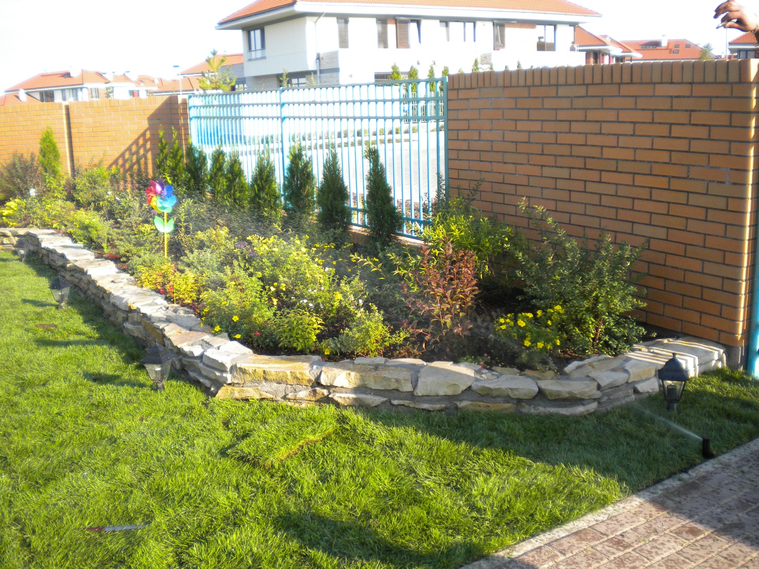 Kaskadowa rabata kwiatowa z kamienną obudową, wyposażona w system nawadniania i ozdobiona kolorowym wiatraczkiem, na tle trawnika, ceglanego muru i niebieskiego ogrodzenia.