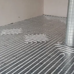 Rozłożona instalacja ogrzewania podłogowego na czarnej folii z nadrukiem, widoczna metalowa rura wentylacyjna w tle, surowy stan wykończenia wnętrza.