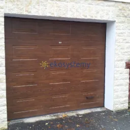 Brązowa segmentowa brama garażowa z widocznym logo firmy, zamontowana w jasnym, kamiennym murze.