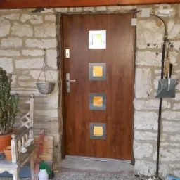 Brązowe drzwi wejściowe z trzema kwadratowymi okienkami z żółtym szkłem, osadzone w kamiennej ścianie, obok ławka z kaktusami w doniczkach i wiszący koszyk.