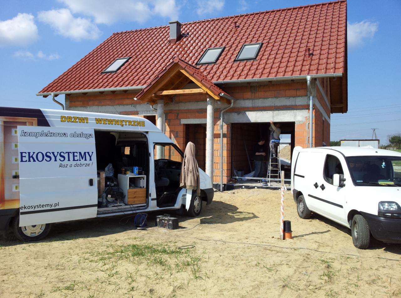 Montaż drzwi wewnętrznych w nowym domu w stanie surowym otwartym, widoczny samochód dostawczy firmy z otwartymi drzwiami i logo, dwóch pracowników na drabinie.