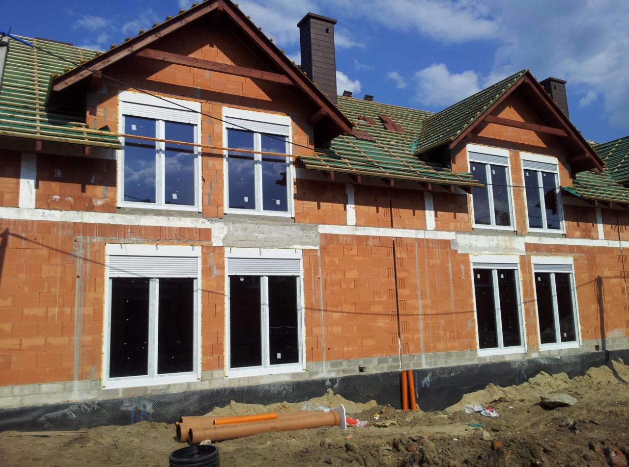 Surowy budynek z cegły z zamontowanymi białymi oknami i częściowo opuszczonymi roletami zewnętrznymi, w tle widoczne rusztowanie dachu i niebieskie niebo.