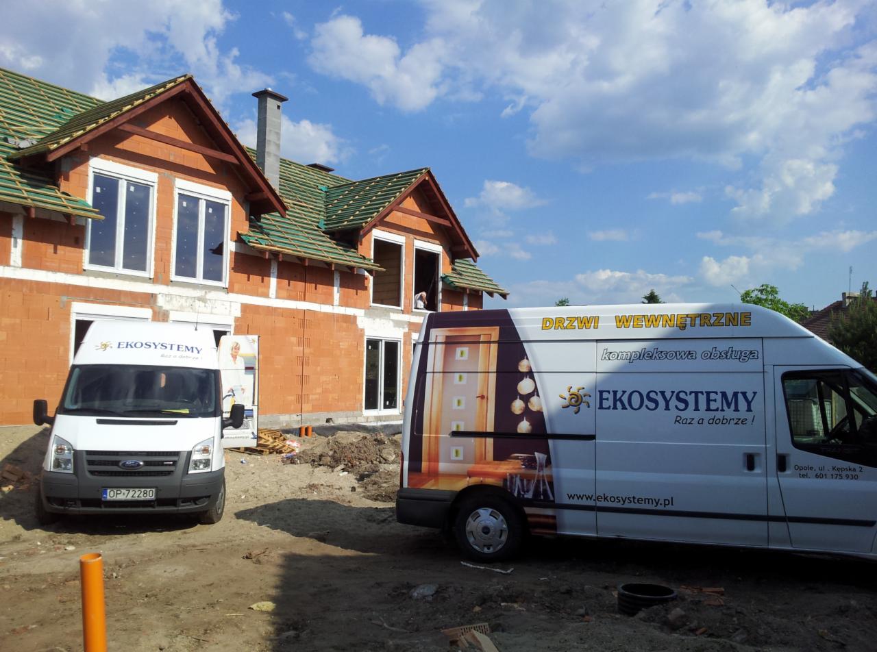 Budowa domu jednorodzinnego w trakcie montażu stolarki okiennej, widoczne dwa busy firmy EkoSystemy z reklamą drzwi wewnętrznych i oknami bez ram w elewacji budynku.