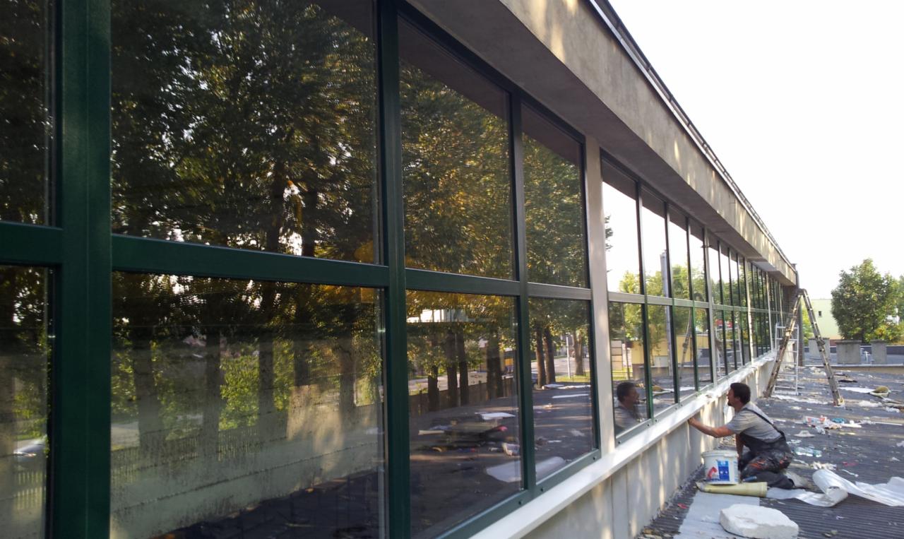 Czyszczenie elewacji budynku z dużymi oknami z zielonymi ramami; mężczyzna w kombinezonie roboczym czyści okno z poziomu dachu, widoczne drzewa odbijające się w szybach.